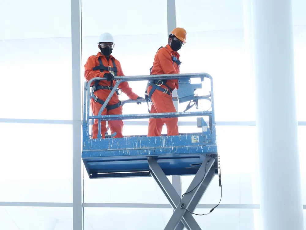 Dos técnicos de Grupo CCN con overol naranja y arnés de seguridad trabajando en una plataforma elevadora de tijera junto a un ventanal